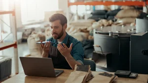 Dueño de negocio hablando por teléfono frente a una laptop en su almacén o planta de producción